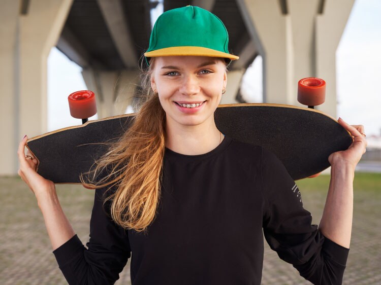 Uśmiechnięta nastoletnia dziewczyna z założonym na ramiona longbordem, w czapce z daszkiem